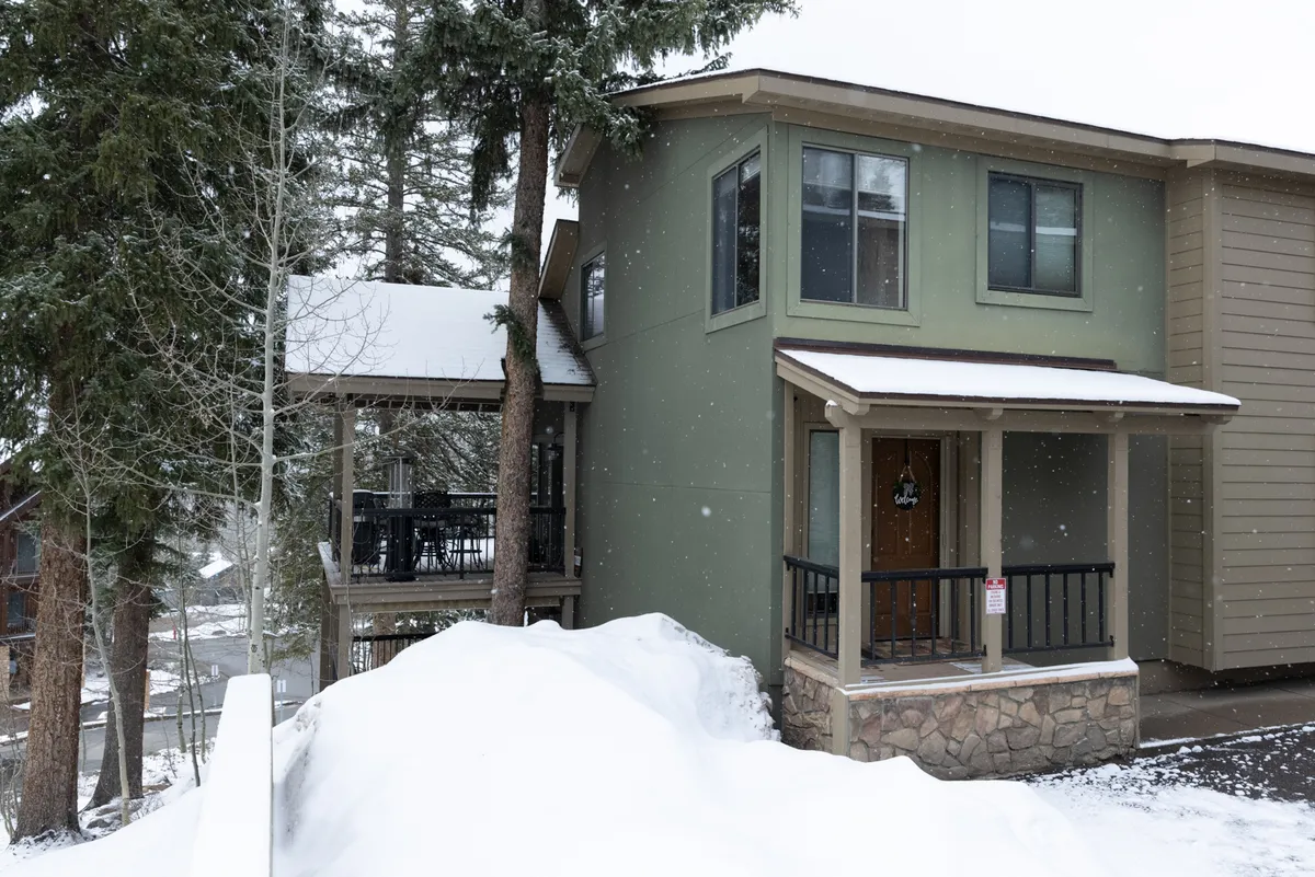 An inviting exterior shot of the property’s front porch, surrounded by snow-laden ground and trees. Large windows reveal a glimpse of the cozy indoors, while the porch offers space for group relaxation and winter fun, such as sipping hot cocoa or watching snowfall together.