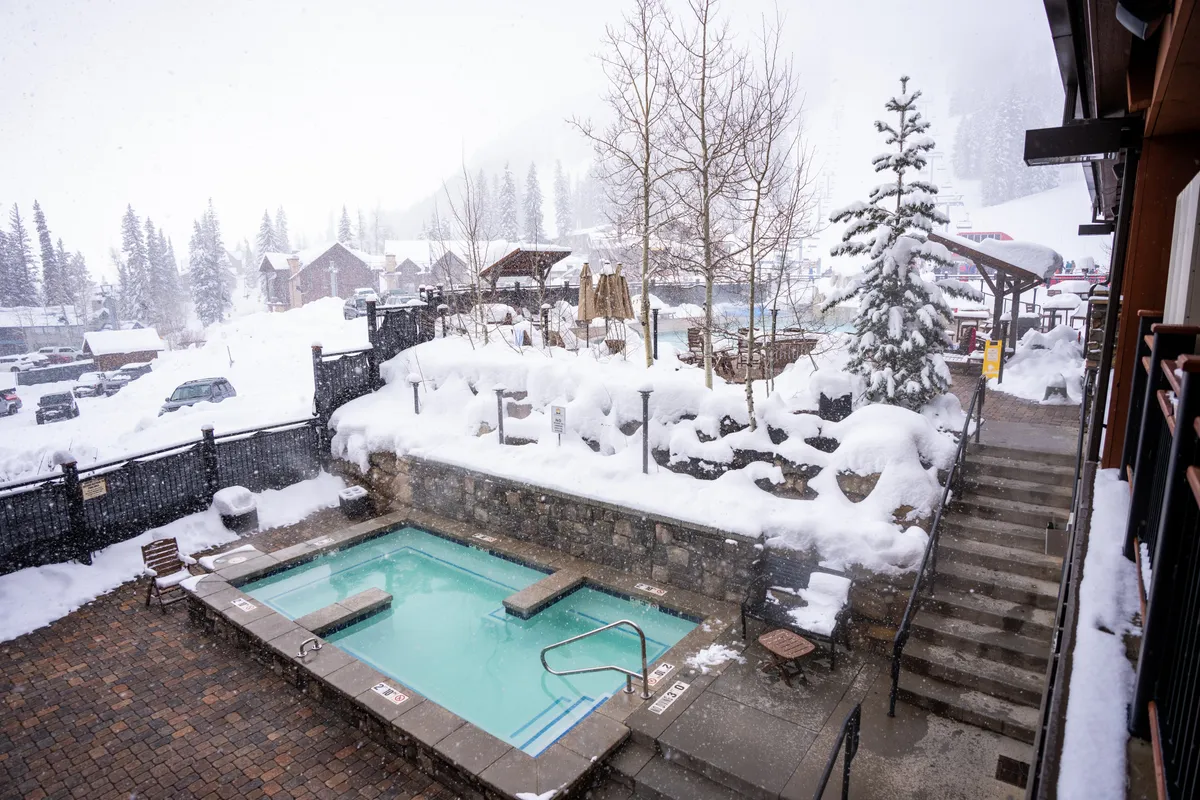 A tranquil outdoor scene featuring a hot tub, a snow-covered landscape, pine trees, and an outdoor seating area. People can relax in the warmth of the hot tub while enjoying the winter scenery or socialize by the fire pit, making it ideal for group interaction after mountain adventures.