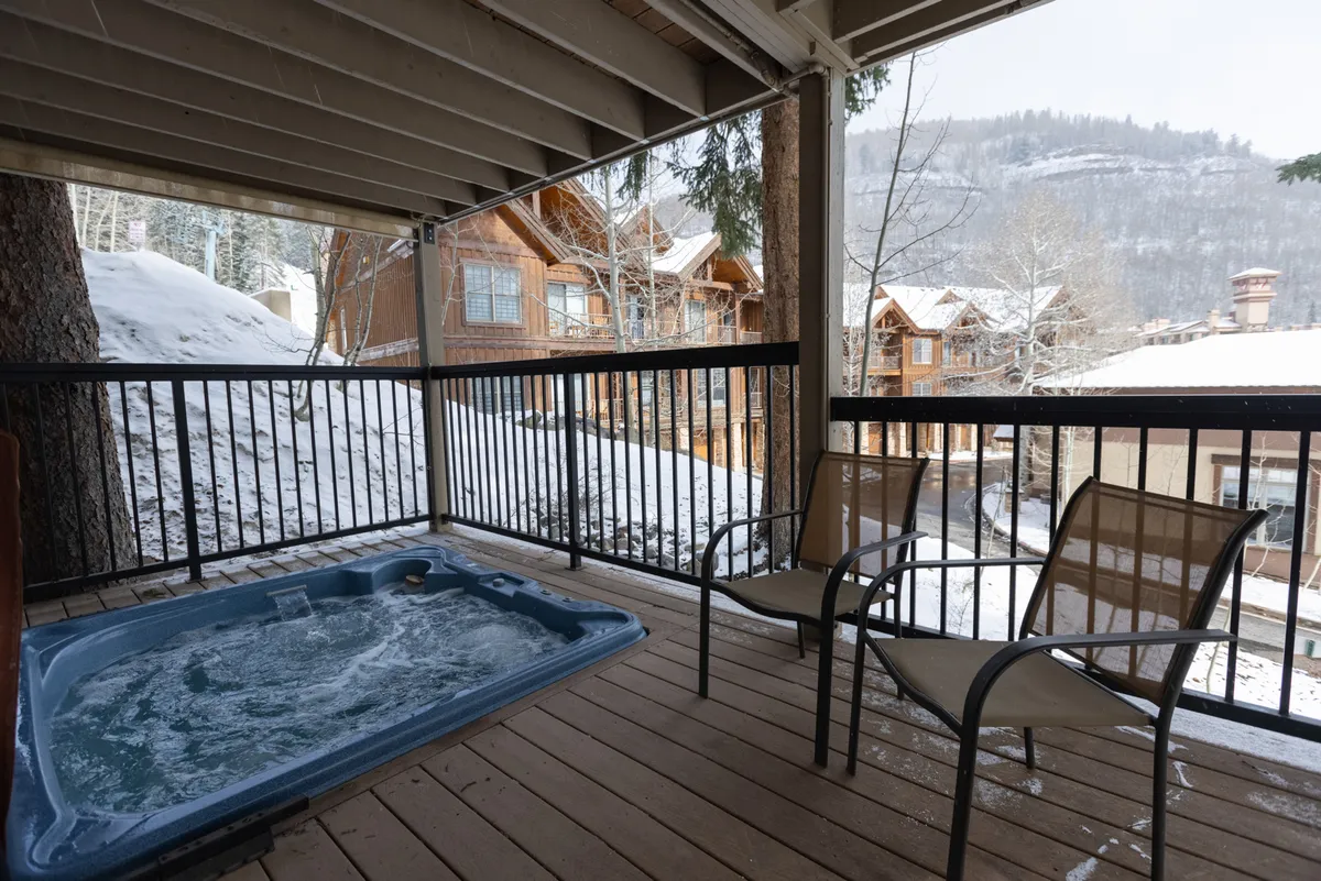 This exterior image features a hot tub on a wooden deck surrounded by trees, nearby cabins, and a backdrop of mountain scenery. The setting is tranquil and inviting, perfect for gatherings or relaxing moments in nature. The deck offers plenty of space for group activities and socializing.