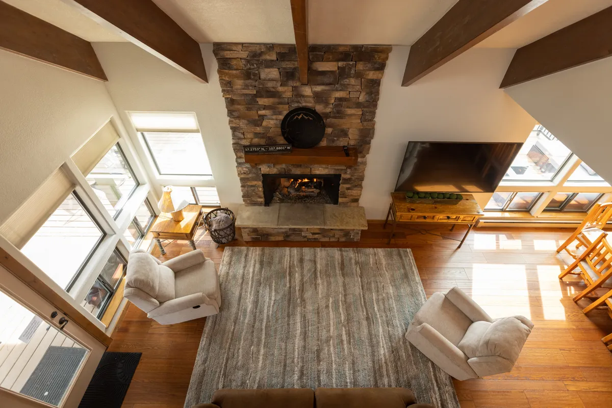 An inviting interior with a rustic stone fireplace, comfortable armchairs, a spacious wooden dining table, and large windows that frame scenic forest views. The room is bathed in natural light, creating a warm, communal atmosphere perfect for relaxation and socializing.