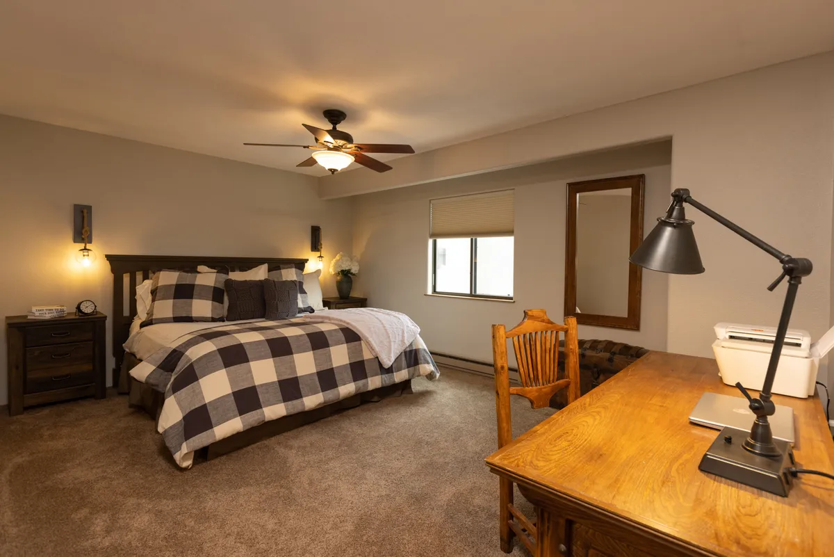 A warmly decorated bedroom with plaid bedding on a cozy bed, a wooden desk for reading or working, and a ceiling fan providing gentle comfort, all contributing to a welcoming environment ideal for personal downtime or productivity.