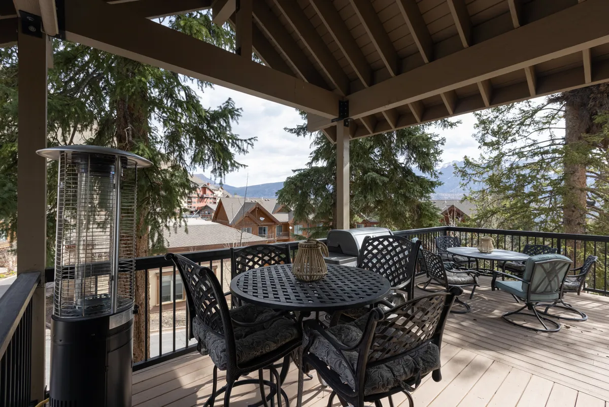 The image features a round outdoor dining table with chairs and additional outdoor seating, set on a spacious deck overlooking a scenic mountain view. Visible heaters suggest the area can be enjoyed year-round, and the inviting layout hints at gatherings and relaxed outdoor meals.