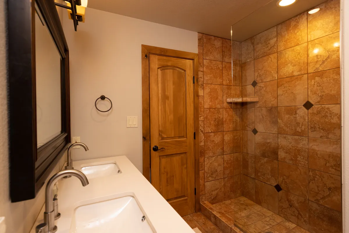 A spacious bathroom featuring a double-sink vanity, a walk-in shower with tiled walls, and a wooden door, offering a calm and inviting environment for relaxation and personal care routines after a long day outdoors.