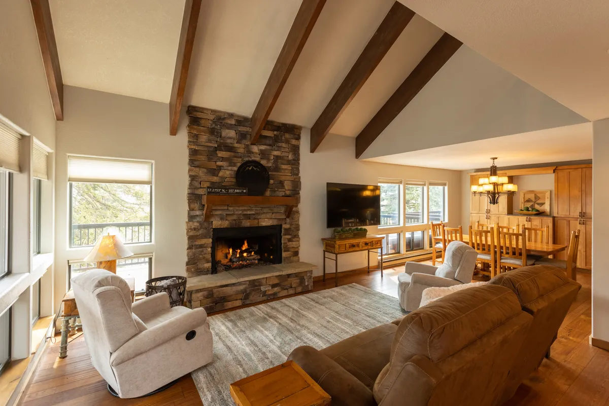 A spacious great room featuring a prominent fireplace surrounded by plush, comfortable seating. The living area is open to a nearby dining space, both brightened by large panoramic windows that offer views of the outdoors. The setting invites gatherings, relaxation, and shared meals, all in a cozy, welcoming atmosphere.