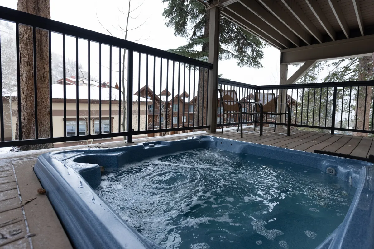 A tranquil outdoor deck scene featuring a hot tub surrounded by deck railings, snow-covered trees, and a wide view of the winter landscape. The setting is perfect for relaxing after a day on the slopes, with space for socializing and unwinding.