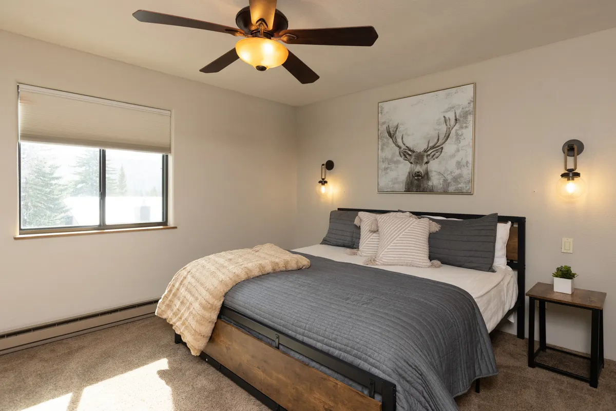 A serene bedroom featuring a comfortable bed with plush linens, tasteful artwork above the headboard, a large window bringing in natural light, and a nightstand with a leafy plant, creating a tranquil atmosphere ideal for relaxation, reading, or unwinding after an active day.