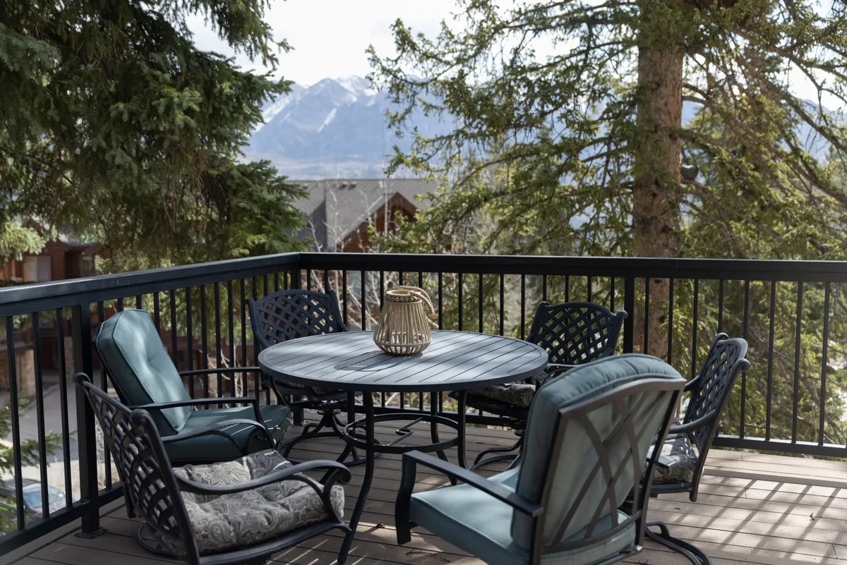 The image shows the exterior wraparound deck of the townhome, featuring a round table set for outdoor dining, a decorative vase centerpiece, lush surrounding trees, and expansive views of the San Juan Mountains. The scene suggests a peaceful, inviting space perfect for gatherings or quiet moments, with the mountain landscape as a stunning backdrop.